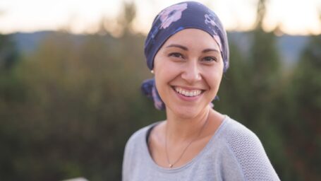 A beautiful young woman wearing a head wrap looks toward the camera and smiles radiantly. She is standing outdoors and there are mountains and trees in the background.